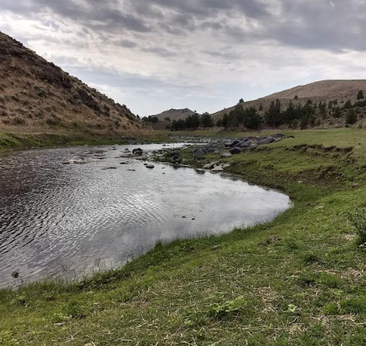 A river flows between grassy banks and hills under a cloudy sky.