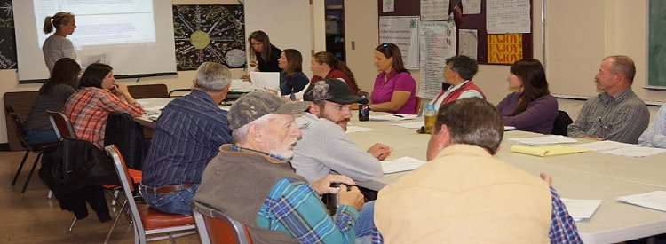 A group of people seated at tables in a meeting room with a presenter standing at the front.