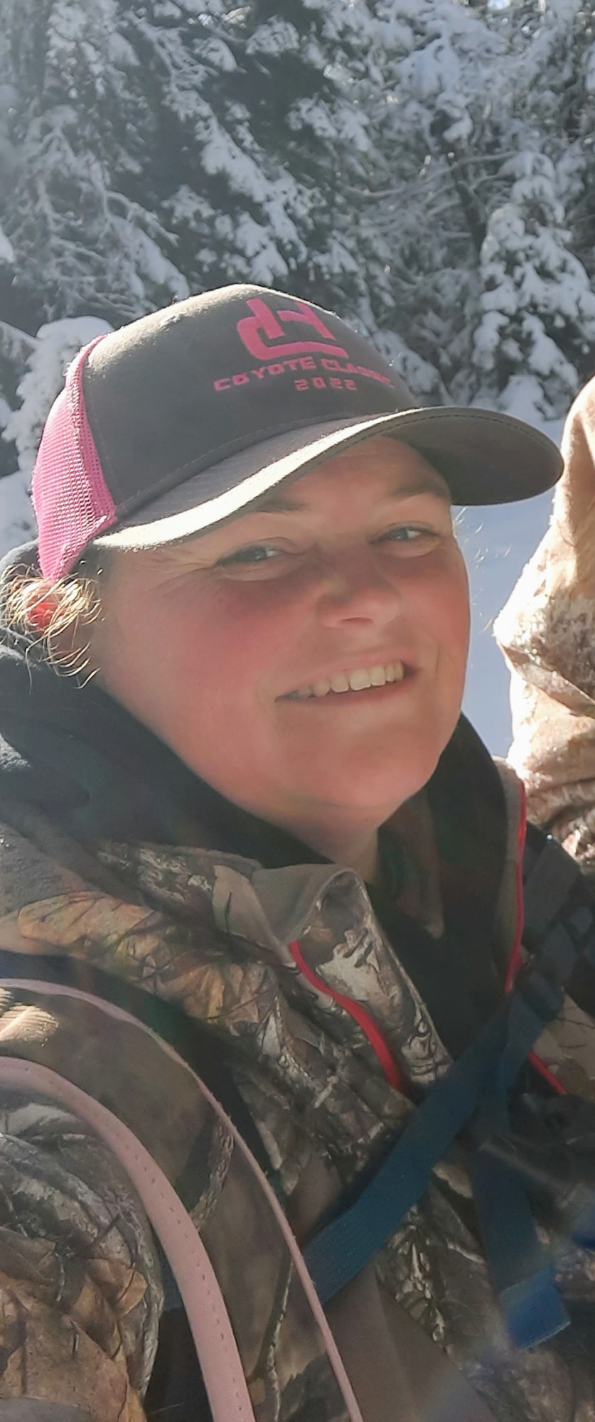 Woman smiling in a woodland setting, wearing a "Coyote Classic 2022" cap and camo jacket.