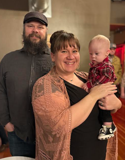 A smiling woman holds a baby in a plaid outfit, with a man in a gray shirt and hat standing behind them. Warm, festive setting.