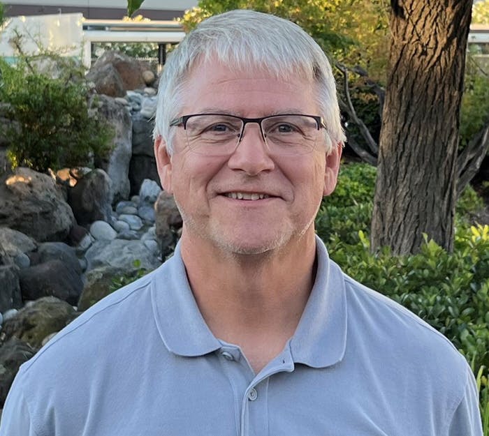 A smiling person with glasses, a grey shirt, with trees and rocks in the background.