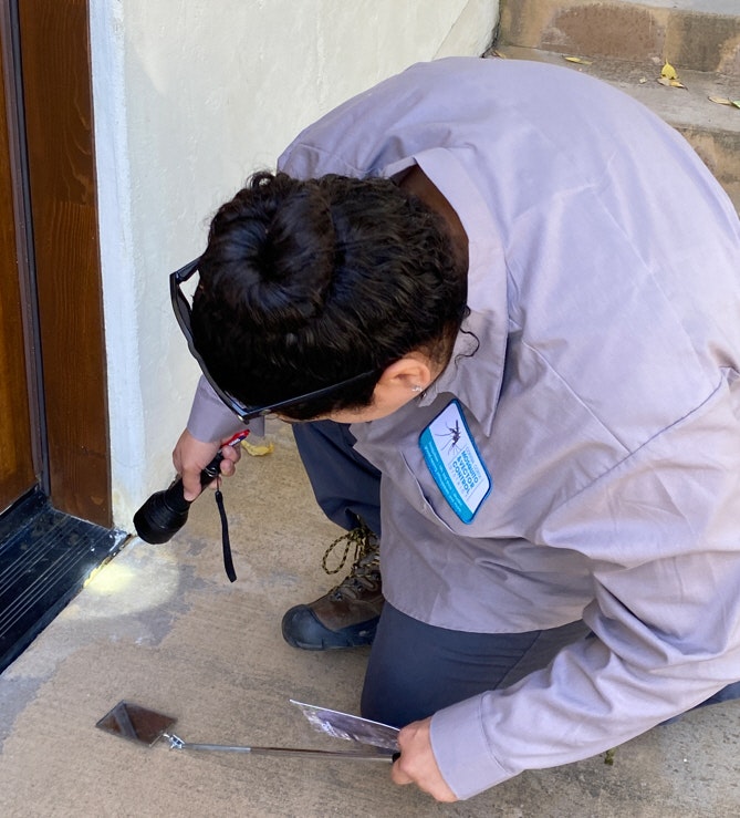 A person is kneeling, holding a flashlight and a mirror, inspecting underneath a doorway.