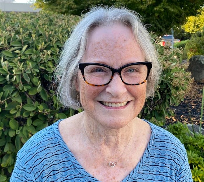 A smiling elderly lady with glasses, wearing a blue top, and a heart necklace, with greenery in the background.