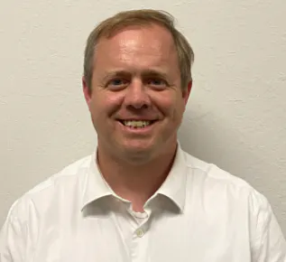 A smiling man wearing a white shirt stands against a light-colored wall.