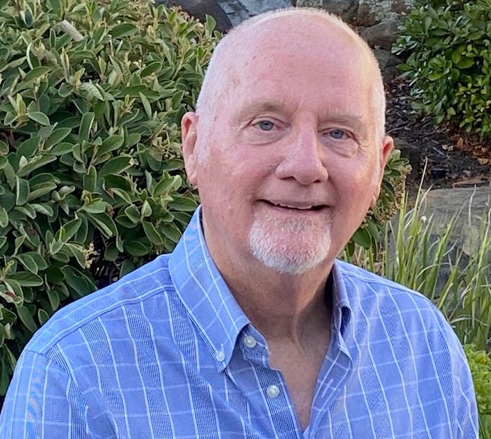A smiling man in a blue checkered shirt with a backdrop of green shrubbery.