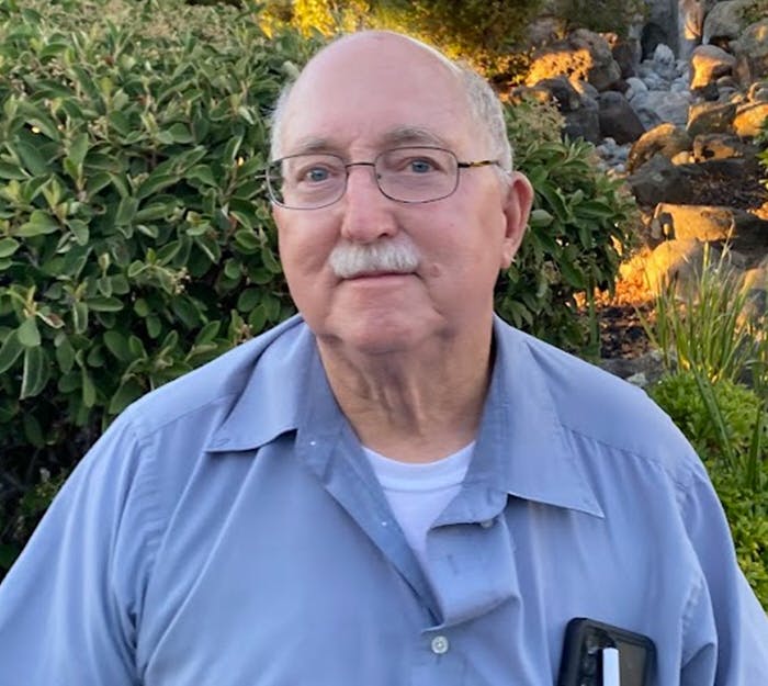 Elderly man with glasses, mustache, and a shirt, standing outdoors with greenery and rocks in the background.