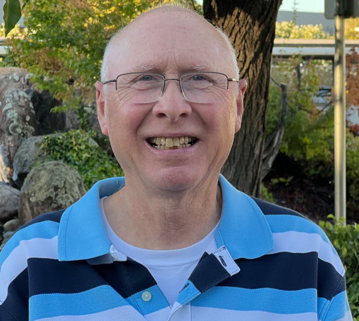 A smiling man wearing glasses and a blue striped polo shirt outdoors.