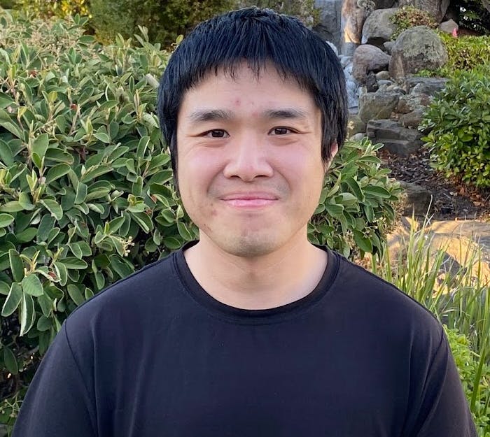 A person smiling in front of greenery with rocks in the background.