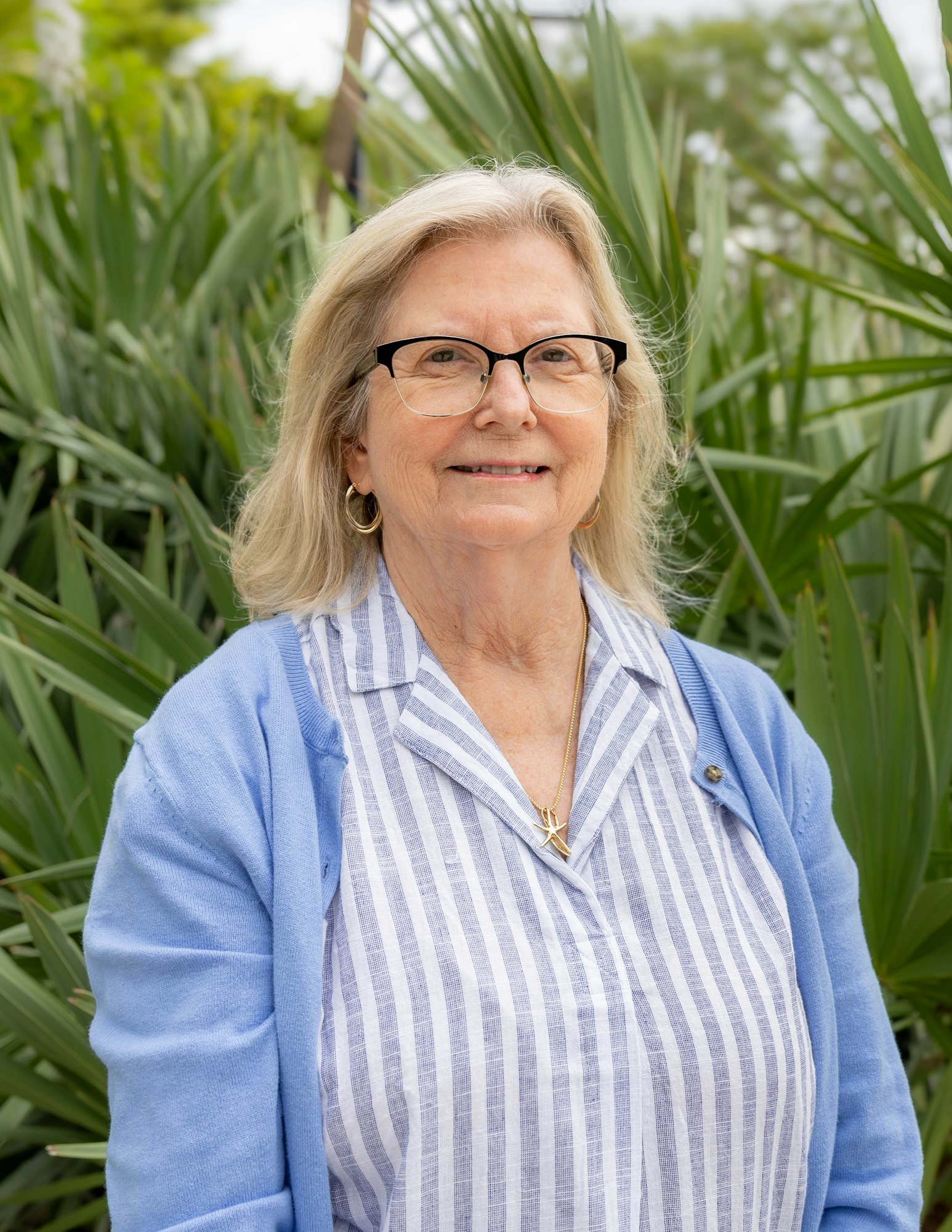A smiling woman with blonde hair, wearing glasses and a blue cardigan, stands in front of green plants.