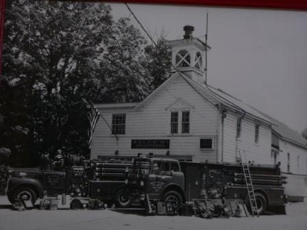 Vintage fire trucks parked in front of an old firehouse with a bell tower, surrounded by trees.