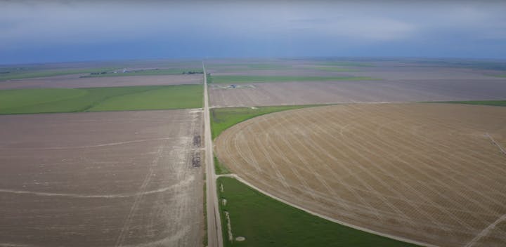 Aerial view of vast farmland with green and brown fields, a road, and a cloudy sky in the background.