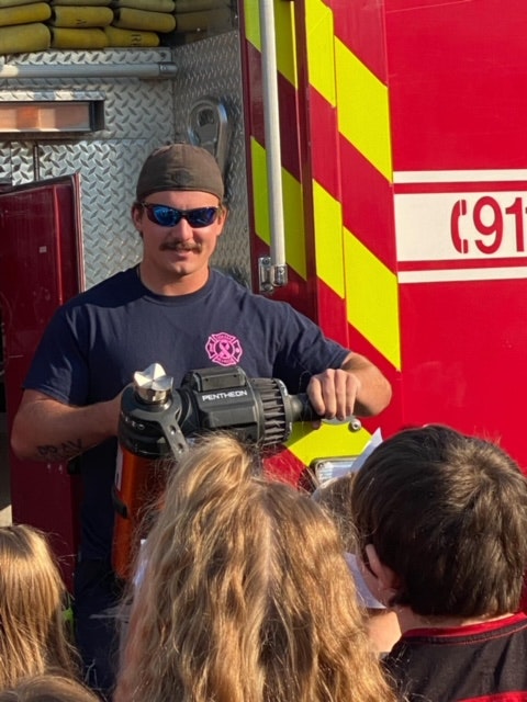 A firefighter is demonstrating equipment to children in front of a fire truck marked "911."