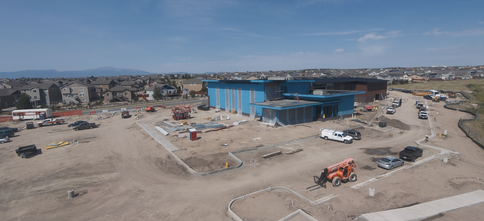 Construction site with a blue building, vehicles, and equipment, surrounded by residential houses under a clear sky.