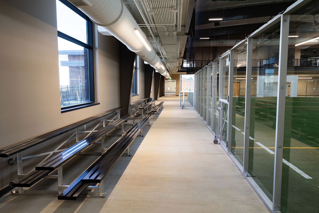 The image shows a spacious indoor corridor with bleacher seating on one side and glass partitions leading to a sports field.