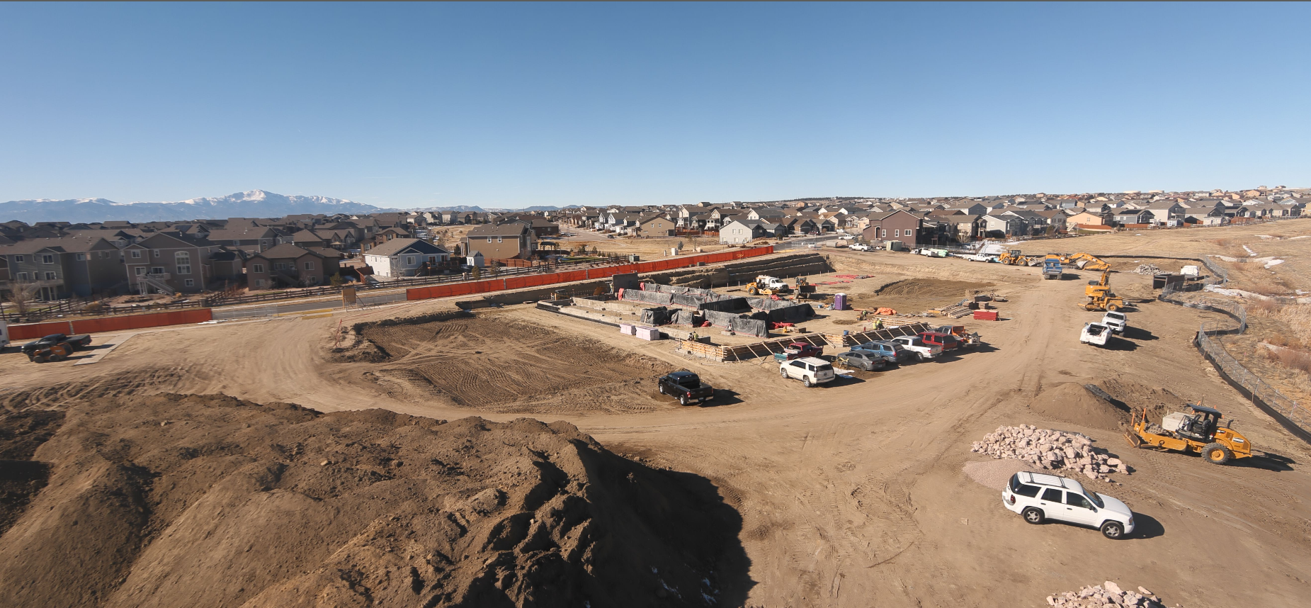 Construction site with vehicles, dirt piles, and a residential neighborhood in the background under a clear blue sky.