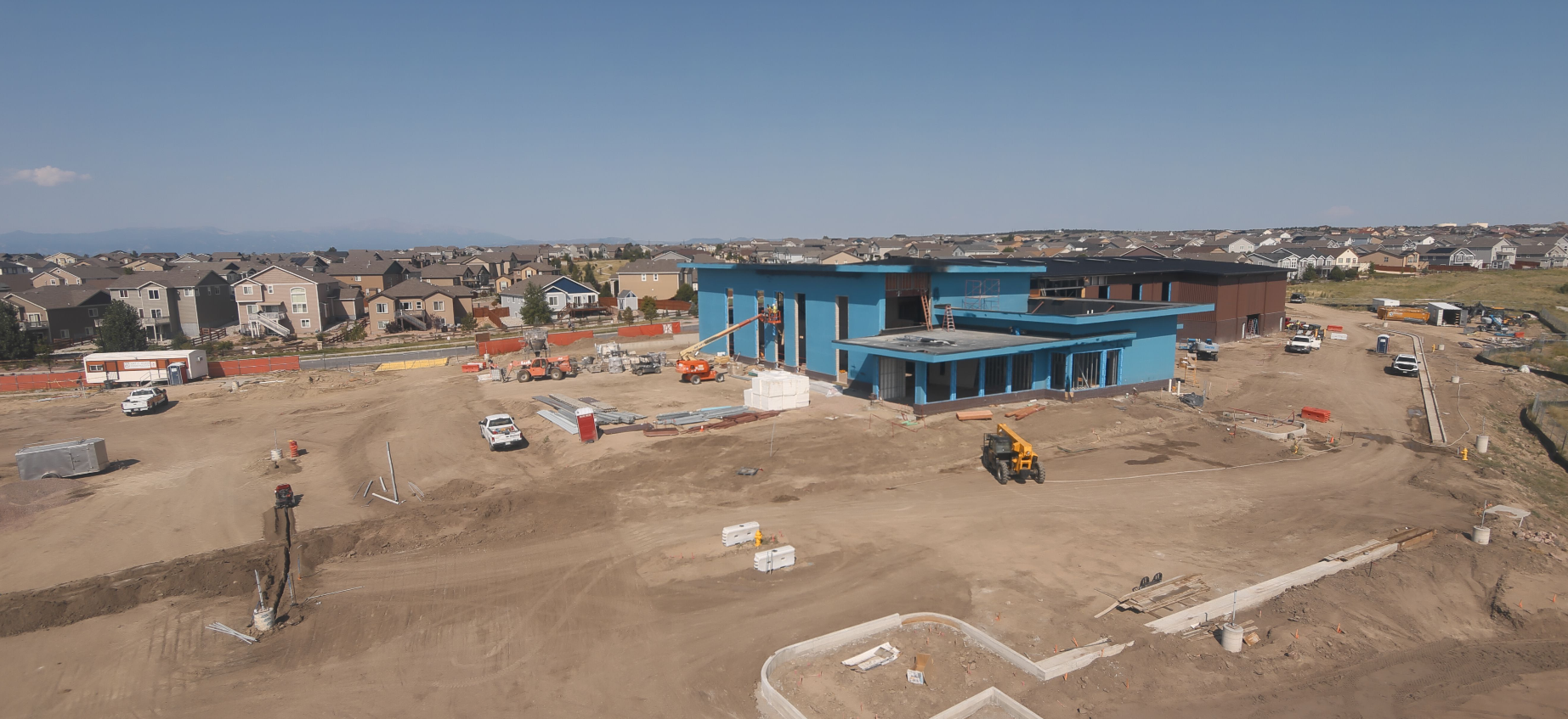 Construction site with a large blue building, machinery, vehicles, and nearby residential houses.