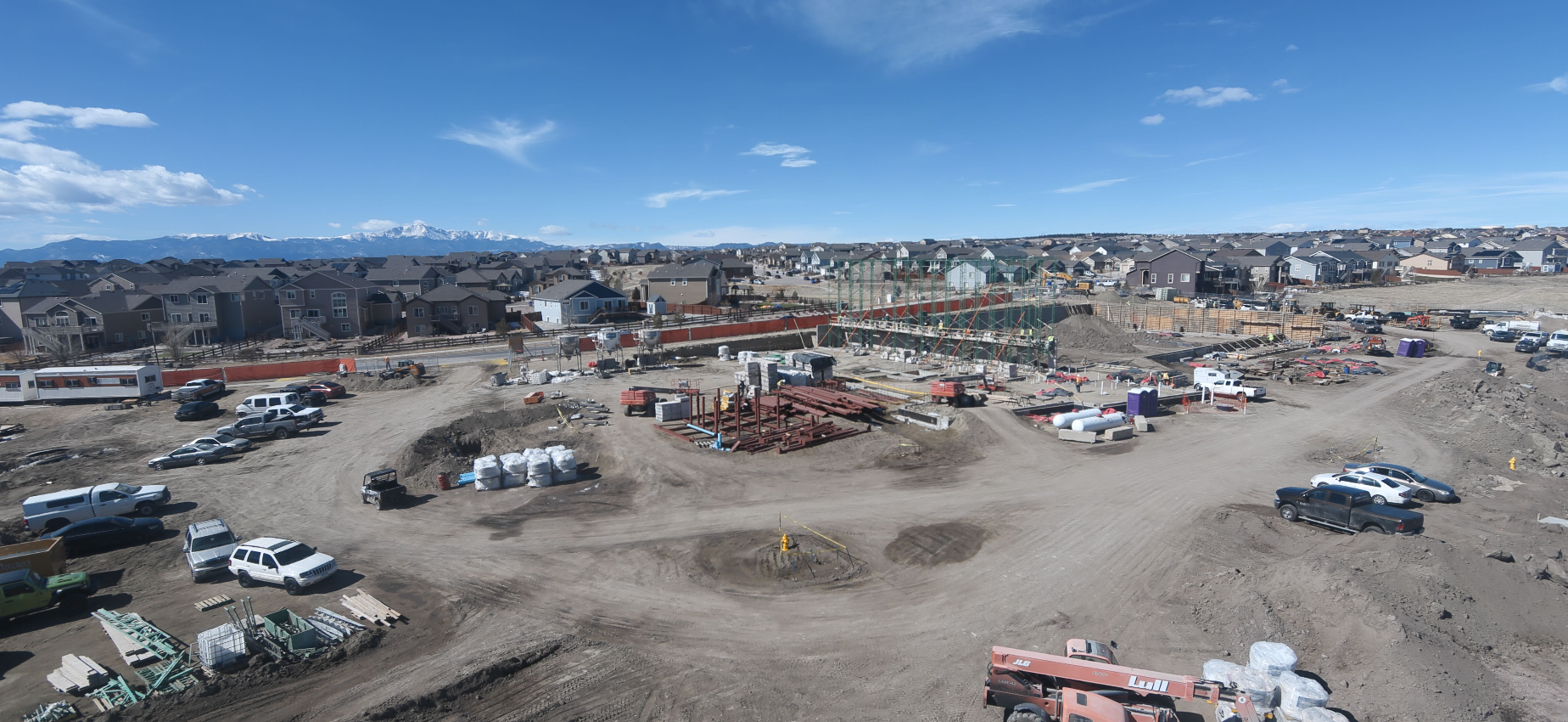 A construction site with vehicles, materials, and scaffolding, surrounded by suburban houses under a clear blue sky.