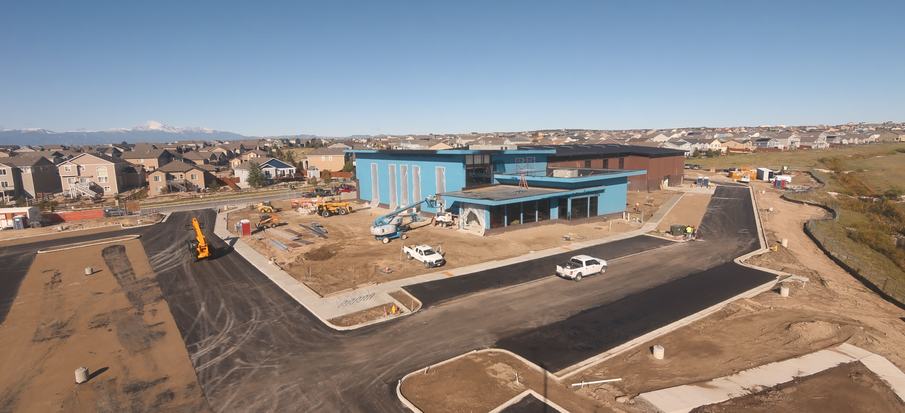 Construction site with a blue building, vehicles, and houses nearby, set against a mountain backdrop.