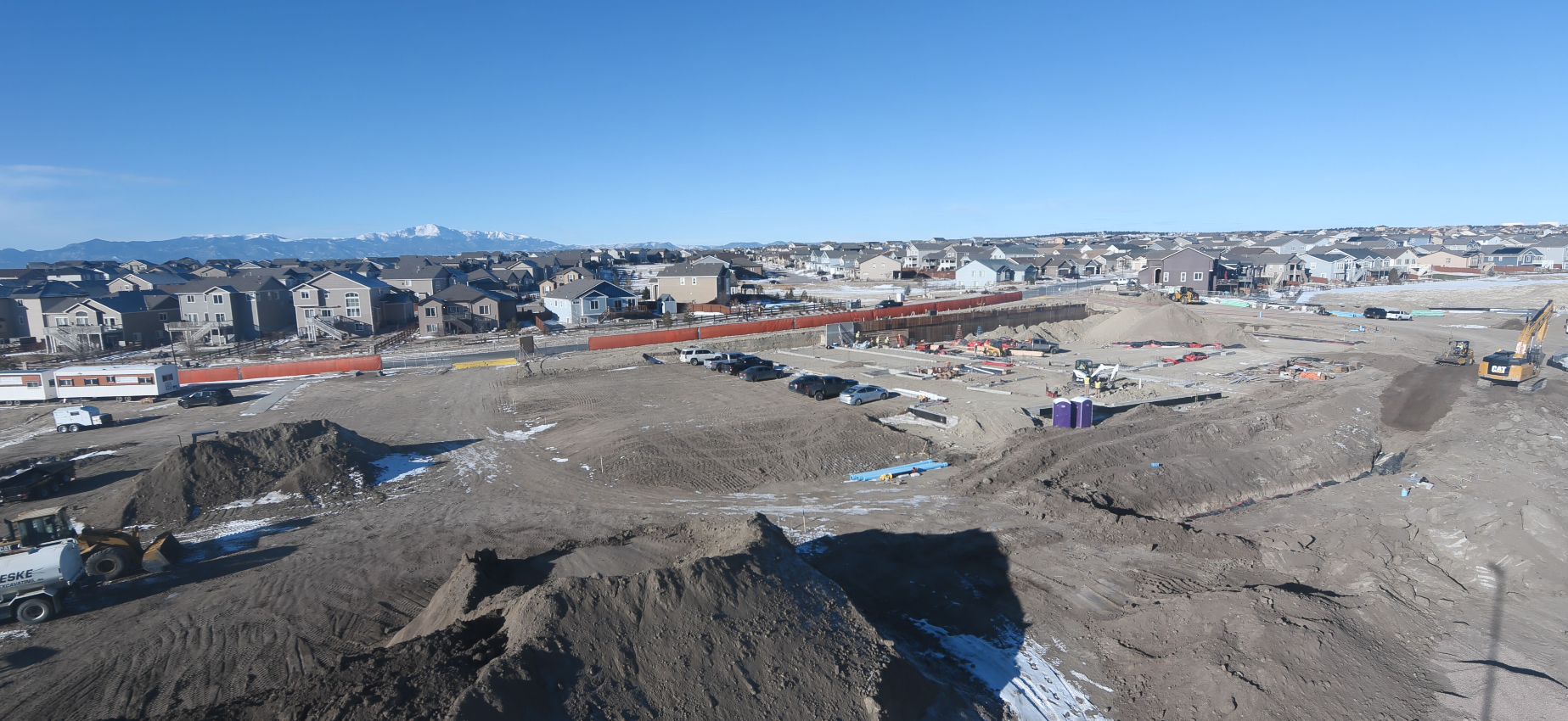 Construction site with heavy machinery, dirt mounds, parked cars, houses, mountains in the background, and a clear blue sky.