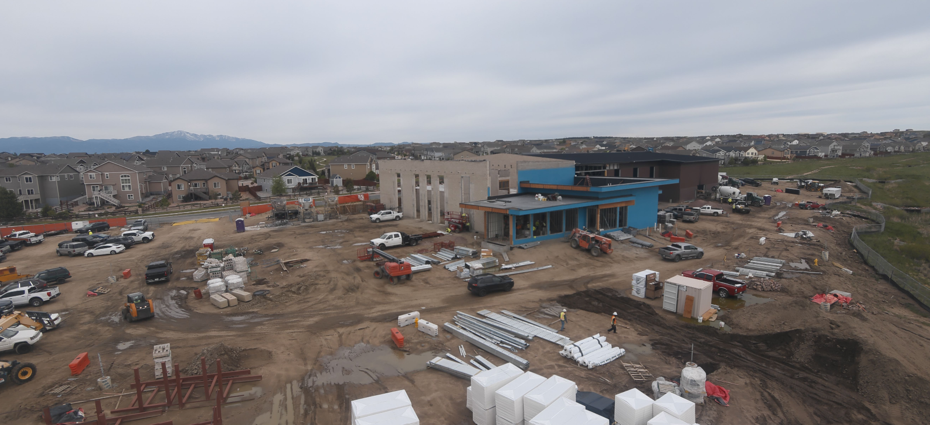 Construction site with vehicles and materials, near residential area, mountain in background.