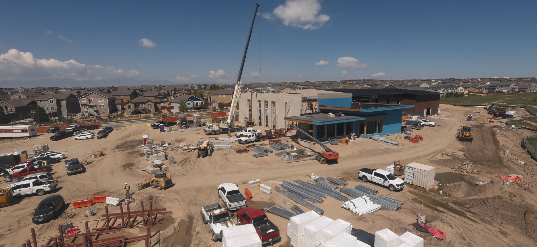 Construction site with various vehicles, equipment, and building materials. Houses and a crane indicate active development.