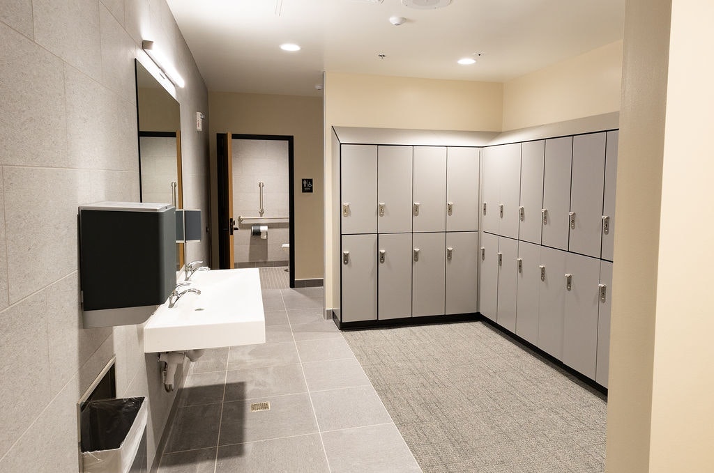 A modern restroom area featuring sinks, a mirror, lockers, and a door leading to another facility. Clean and well-lit design.