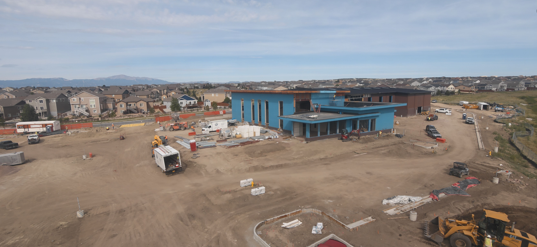 Construction site with a blue building, vehicles, and equipment, surrounded by houses and mountains in the background.