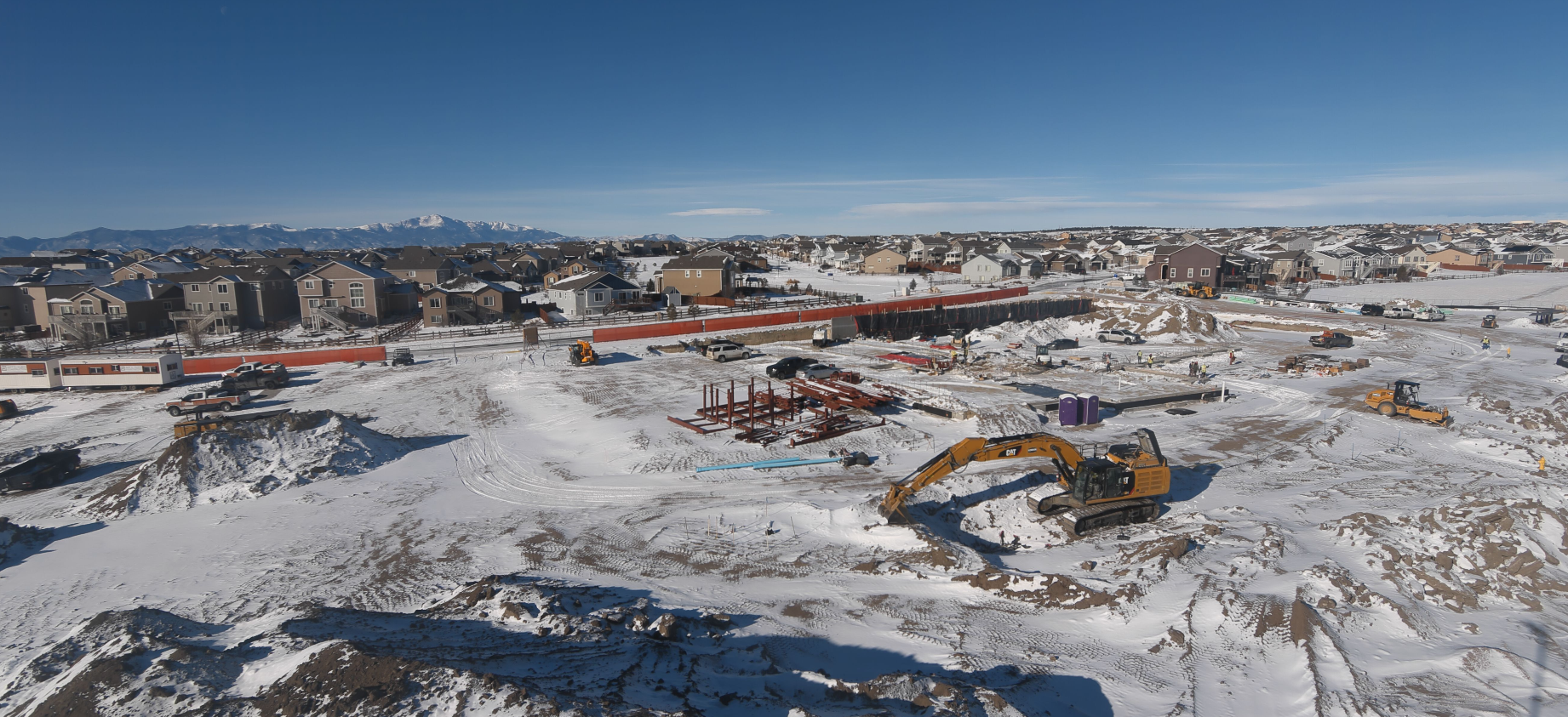 Snowy construction site with machinery, residential houses, and distant mountains under a clear blue sky.