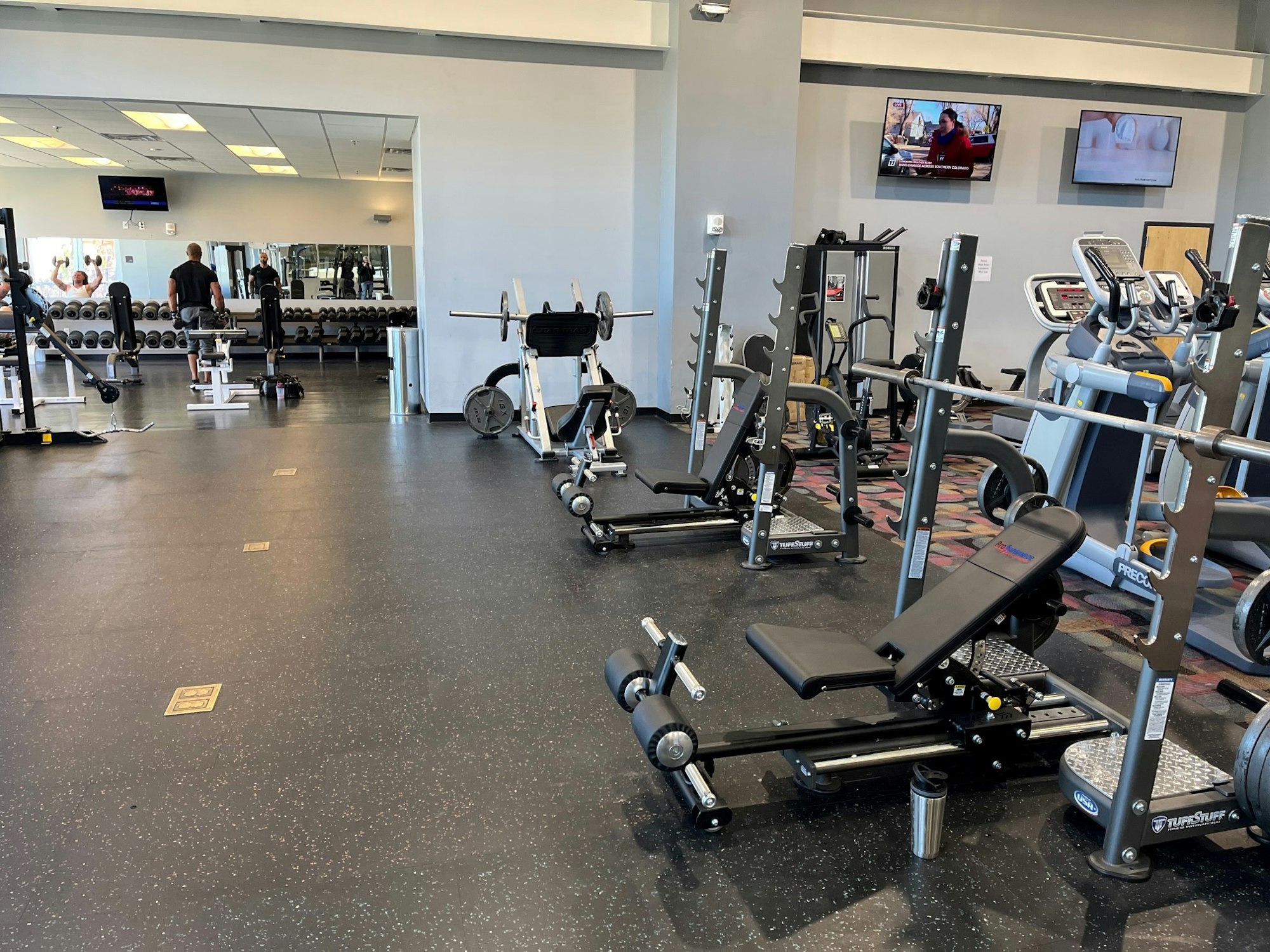 A well-equipped gym featuring weight machines, dumbbells, and cardio equipment, with mirrors and TVs in view.