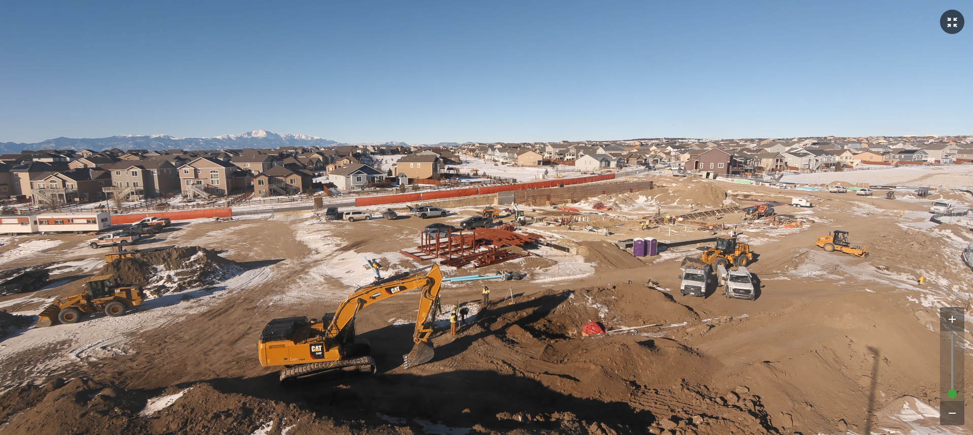 Construction site with machinery, dirt piles, and workers; residential neighborhood and mountains in the background.