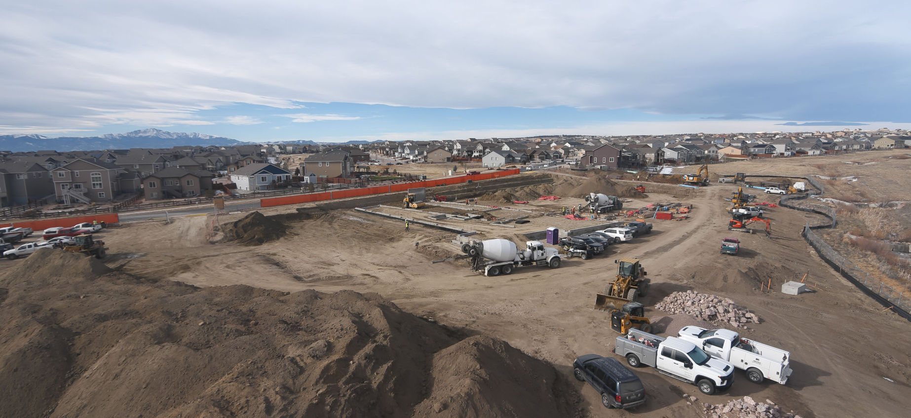 Construction site with vehicles, heavy equipment, and workers. Residential neighborhood and mountains in the background.