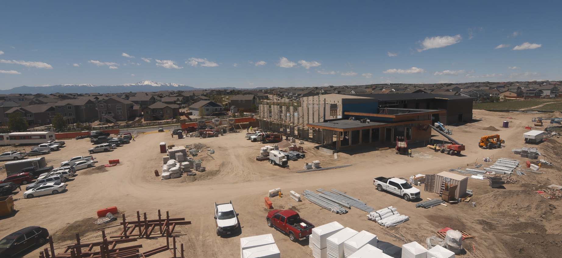 Construction site with vehicles, building materials, and workers. Houses and mountains in the background under a clear sky.