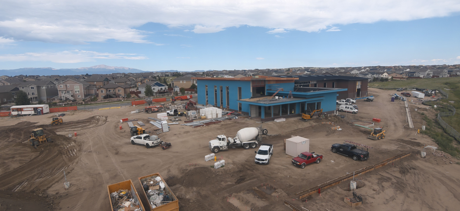 Construction site with trucks and machinery, surrounded by residential houses and a blue building in progress.