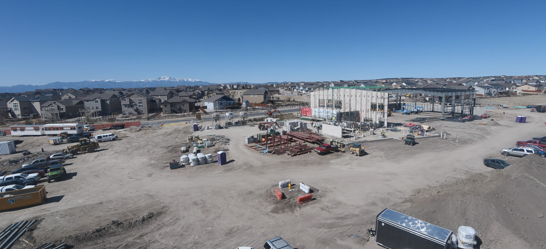 Construction site with workers, vehicles, and building materials. Houses and mountains in the background under a clear blue sky.