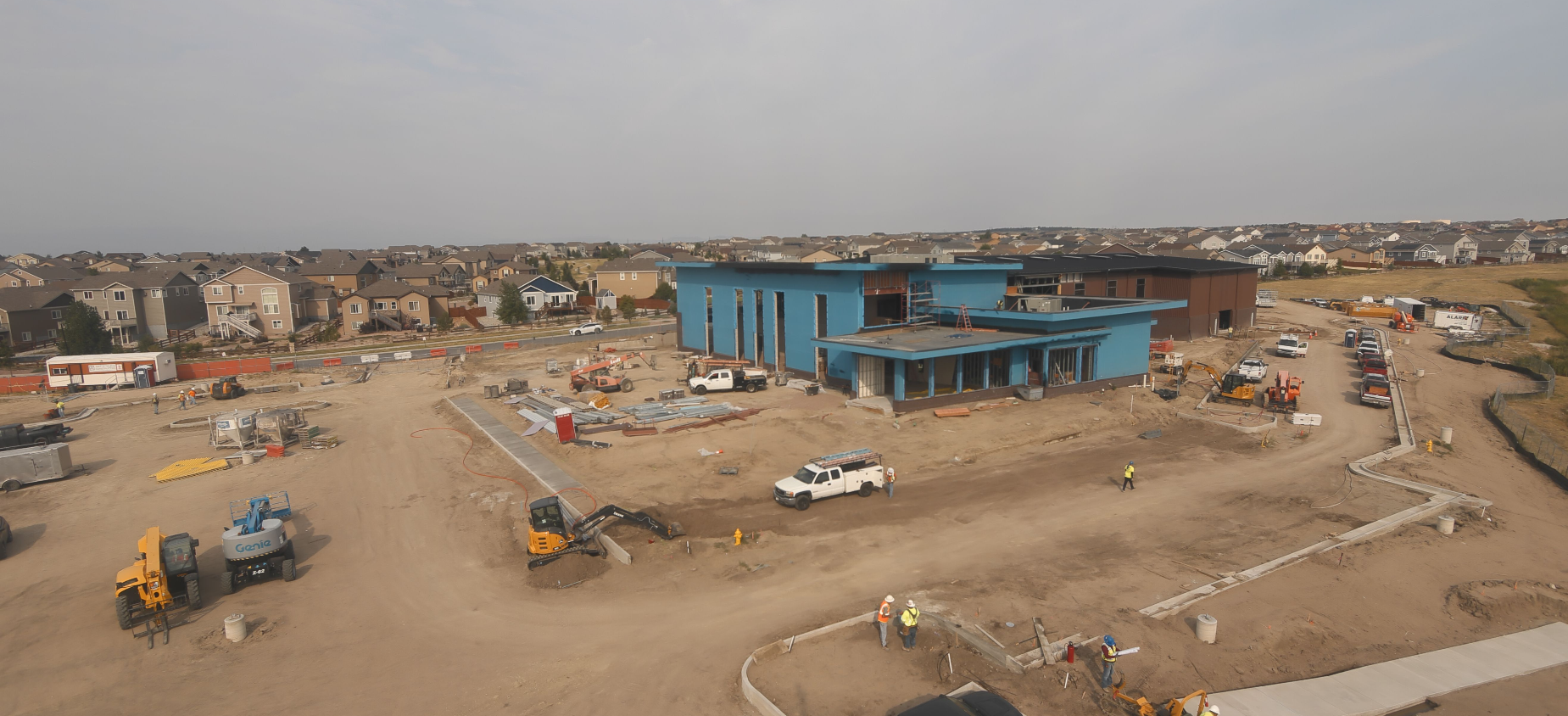 Construction site with a large building, vehicles, and workers, surrounded by houses in the background.