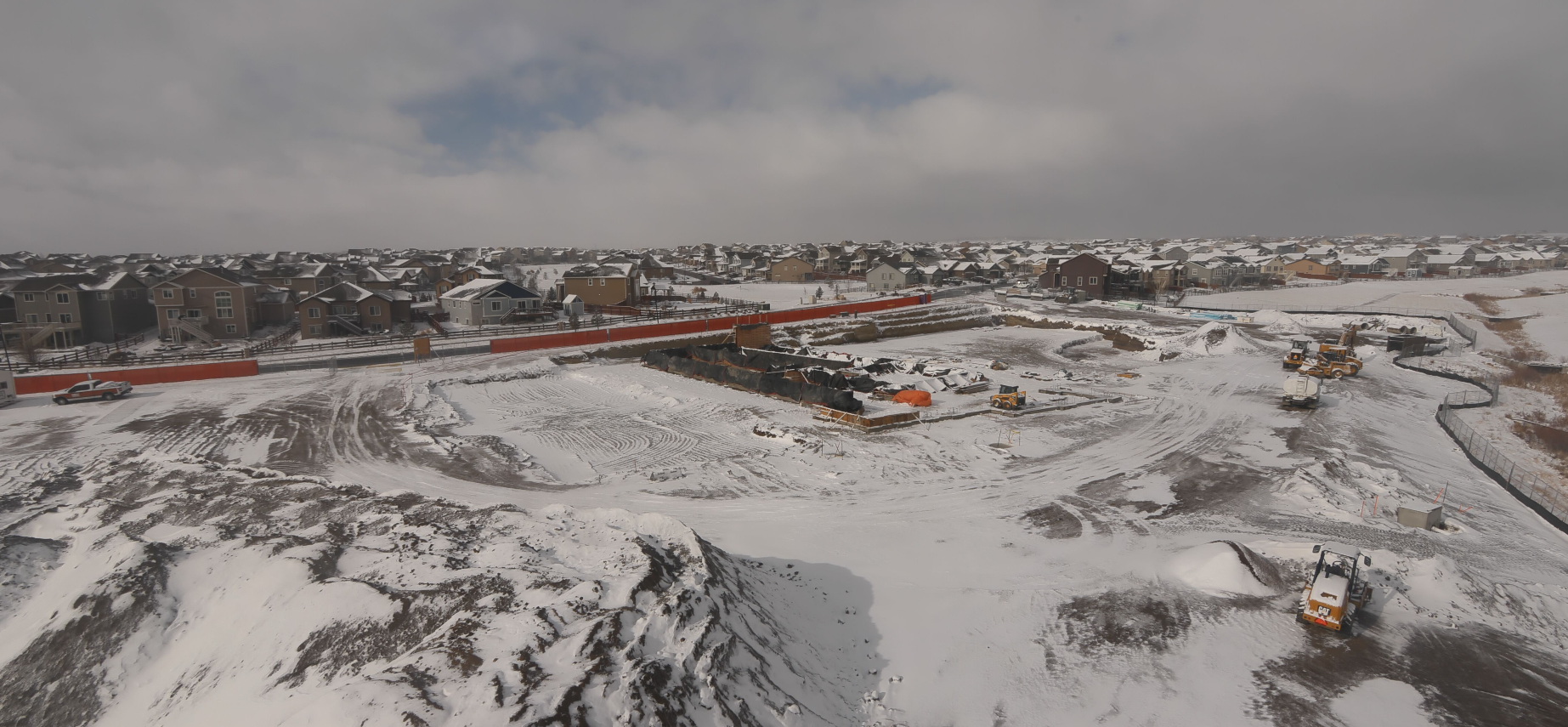 Snowy construction site with heavy machinery, surrounded by residential houses.