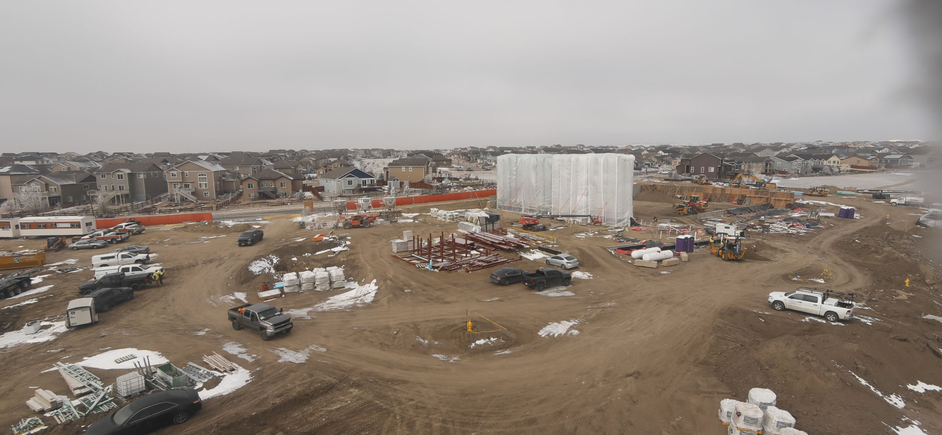 A construction site with vehicles, building materials, and houses in the background, under a cloudy sky.