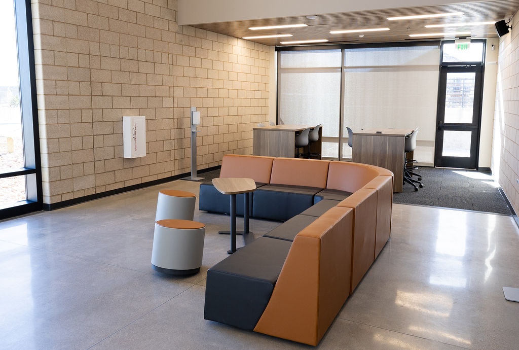 A modern interior space features a curved couch, small tables, and workstations with a large window and natural light.