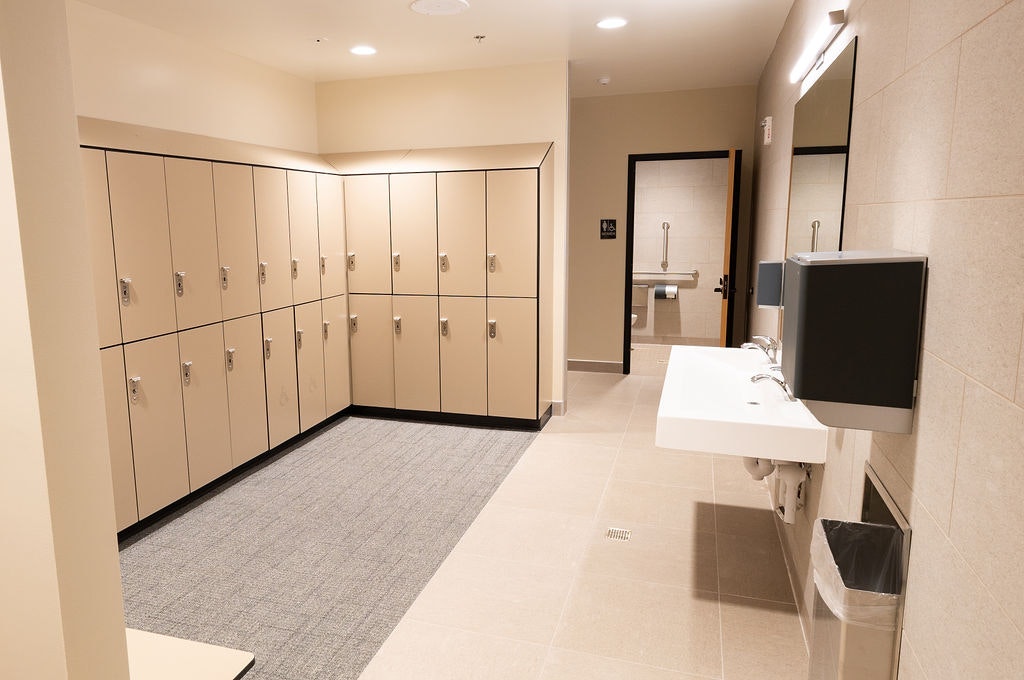 The image shows a modern locker room with beige lockers, sinks, and restroom facilities. It appears clean and well-maintained.