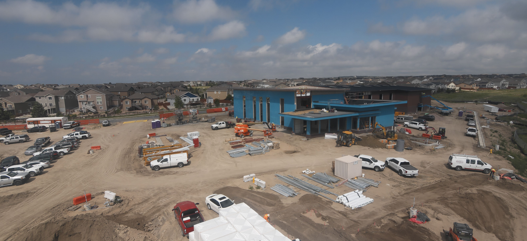 Construction site with a blue building, vehicles, construction materials, and equipment, surrounded by houses under a partly cloudy sky.