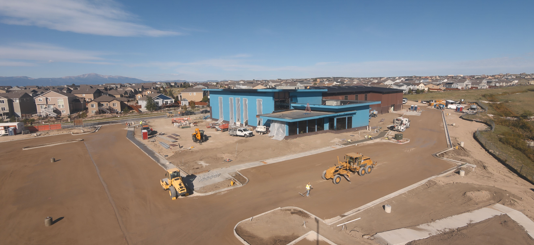 Construction site with a blue building, machinery, and workers, surrounded by residential houses under a clear blue sky.