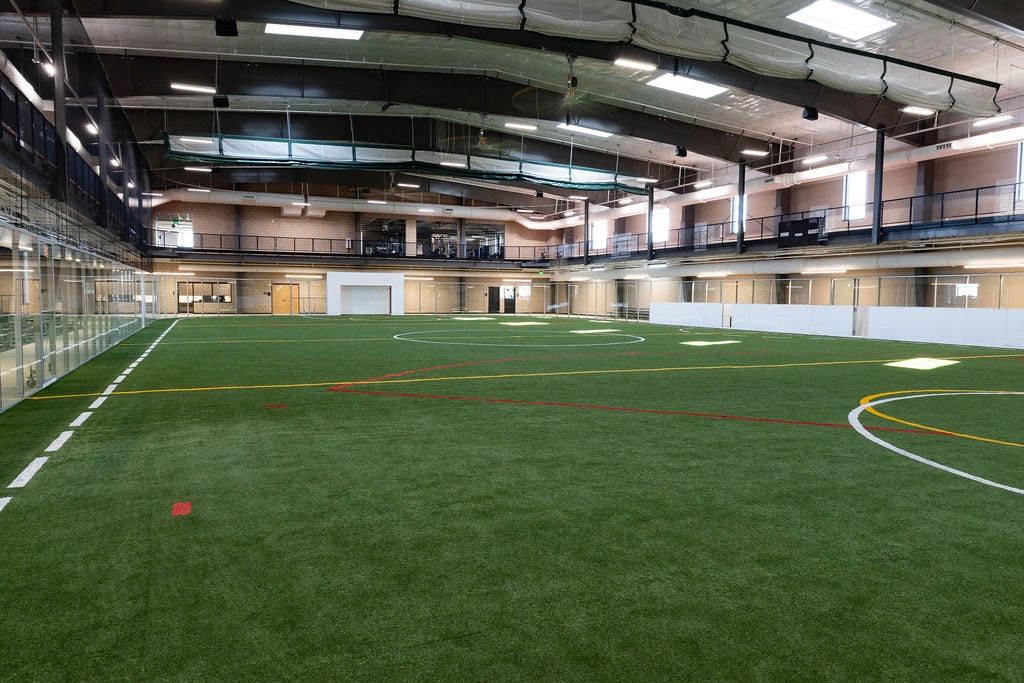 An indoor sports facility featuring a large artificial turf field, surrounded by viewing areas and amenities.