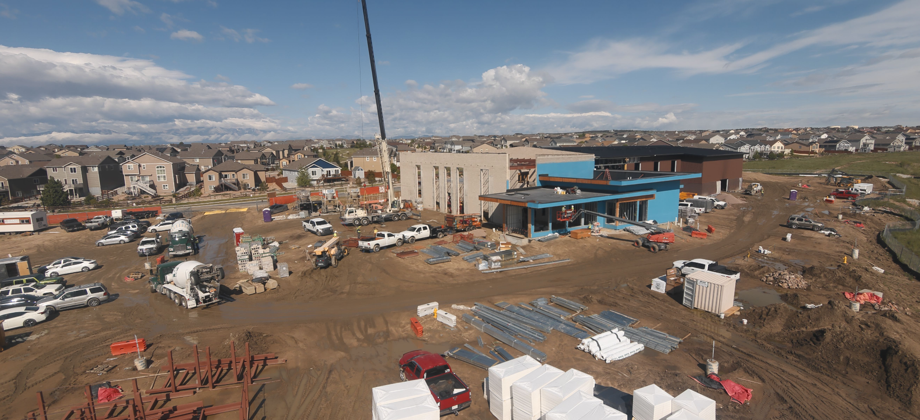 Construction site with vehicles, materials, and buildings in progress, surrounded by residential houses under a partly cloudy sky.