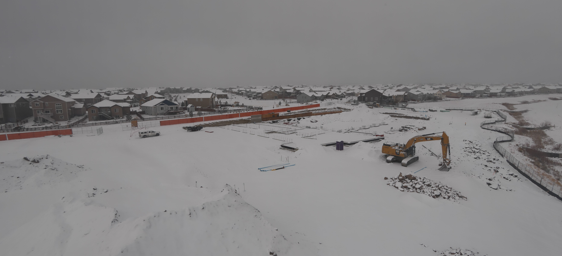 Snow-covered construction site with heavy machinery, near a residential area.