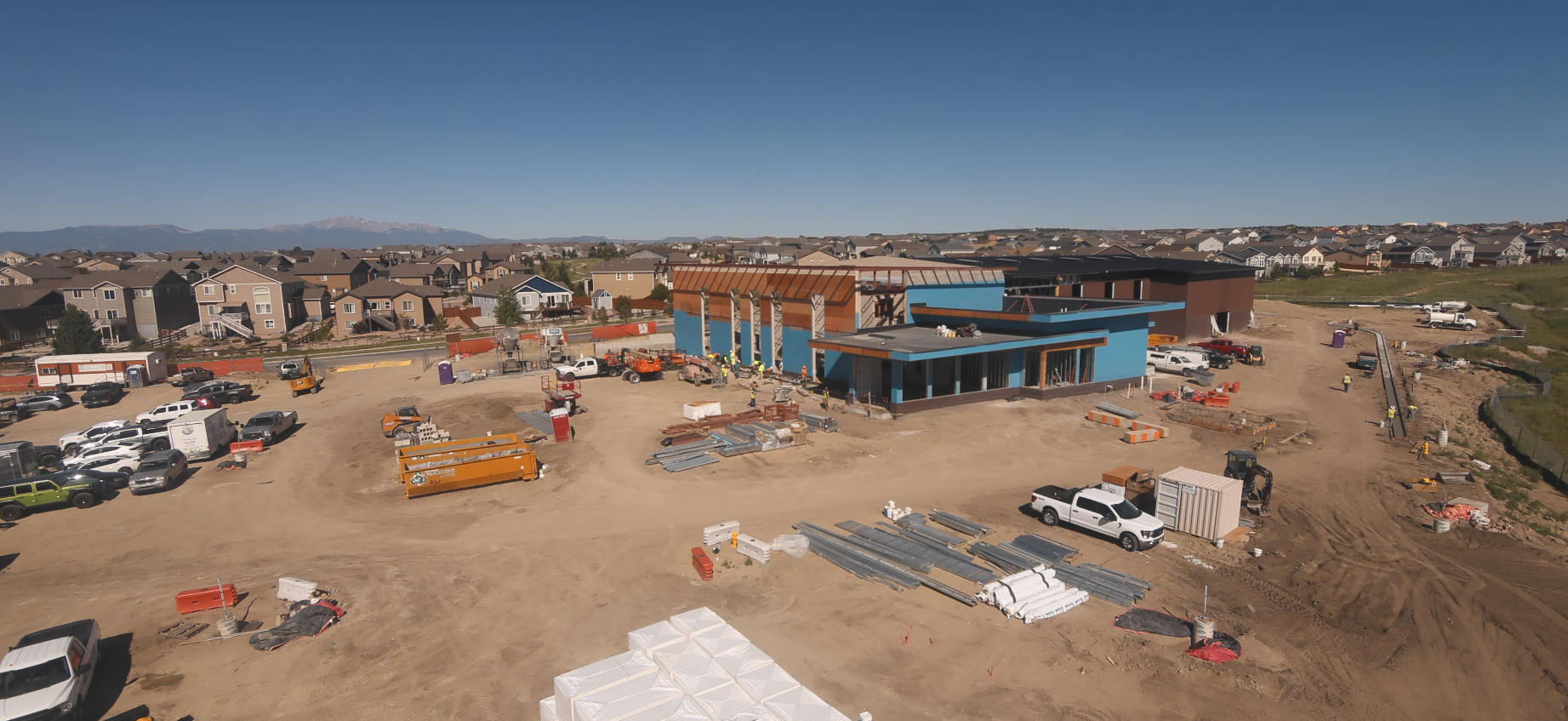 Construction site with machinery, building materials, workers, and nearby residential area under a clear blue sky.