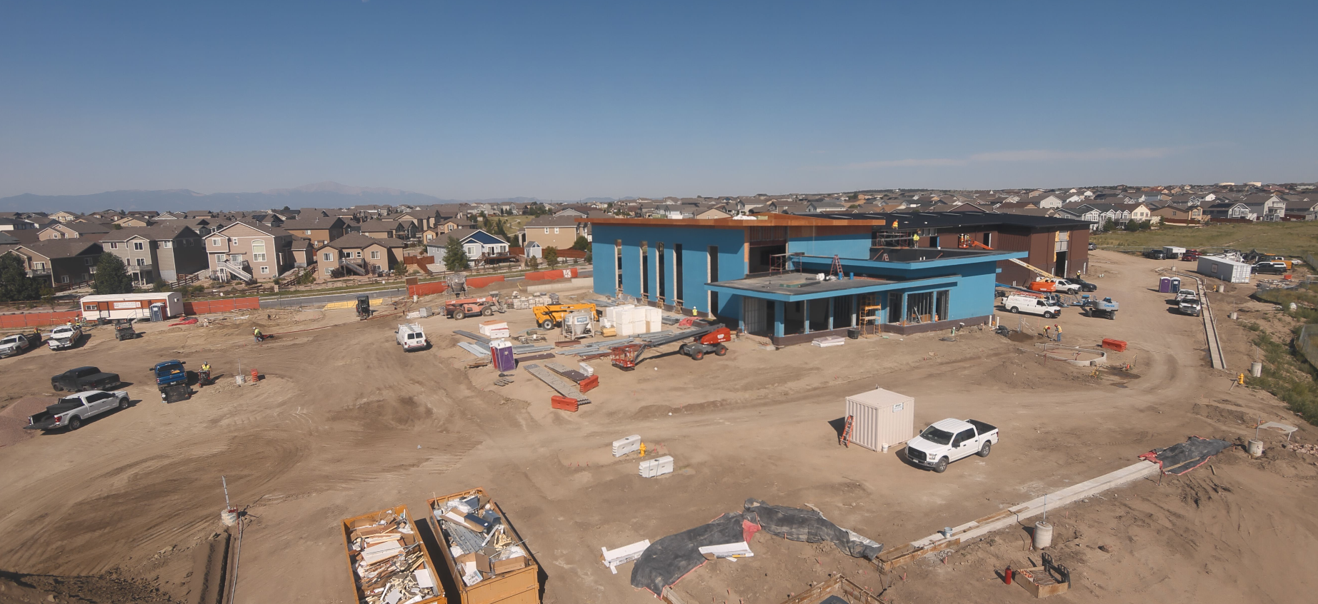 A construction site with a blue building, vehicles, equipment, and nearby houses in a residential area.