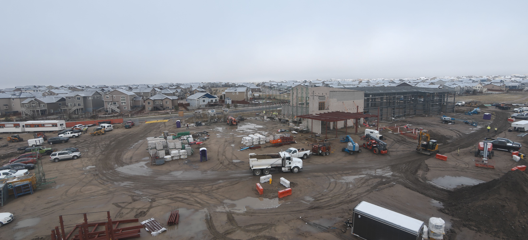 Construction site with machinery, vehicles, and materials; houses in the background under a cloudy sky.