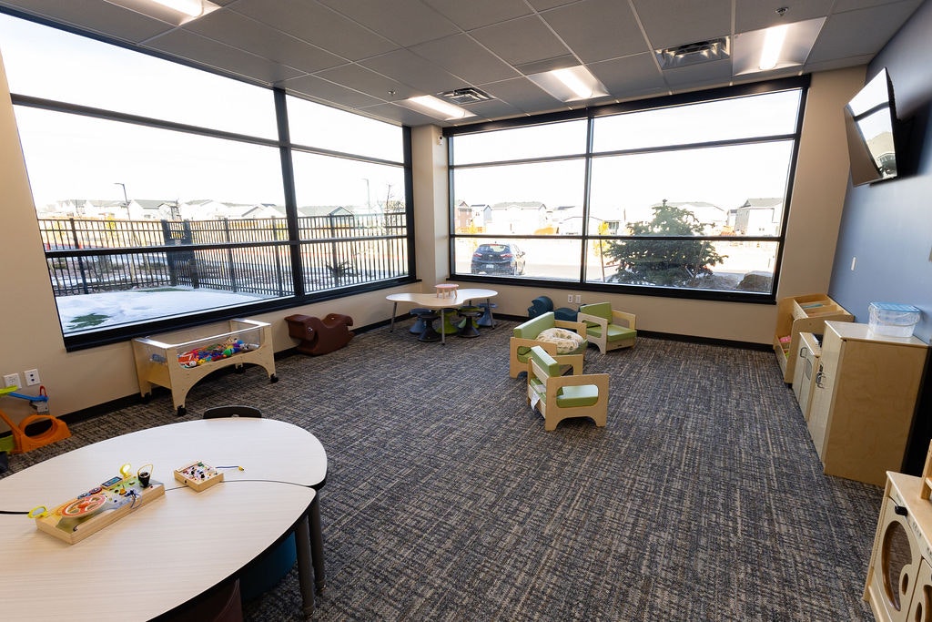A bright, spacious playroom with tables, toys, and seating, featuring large windows overlooking a fenced area.