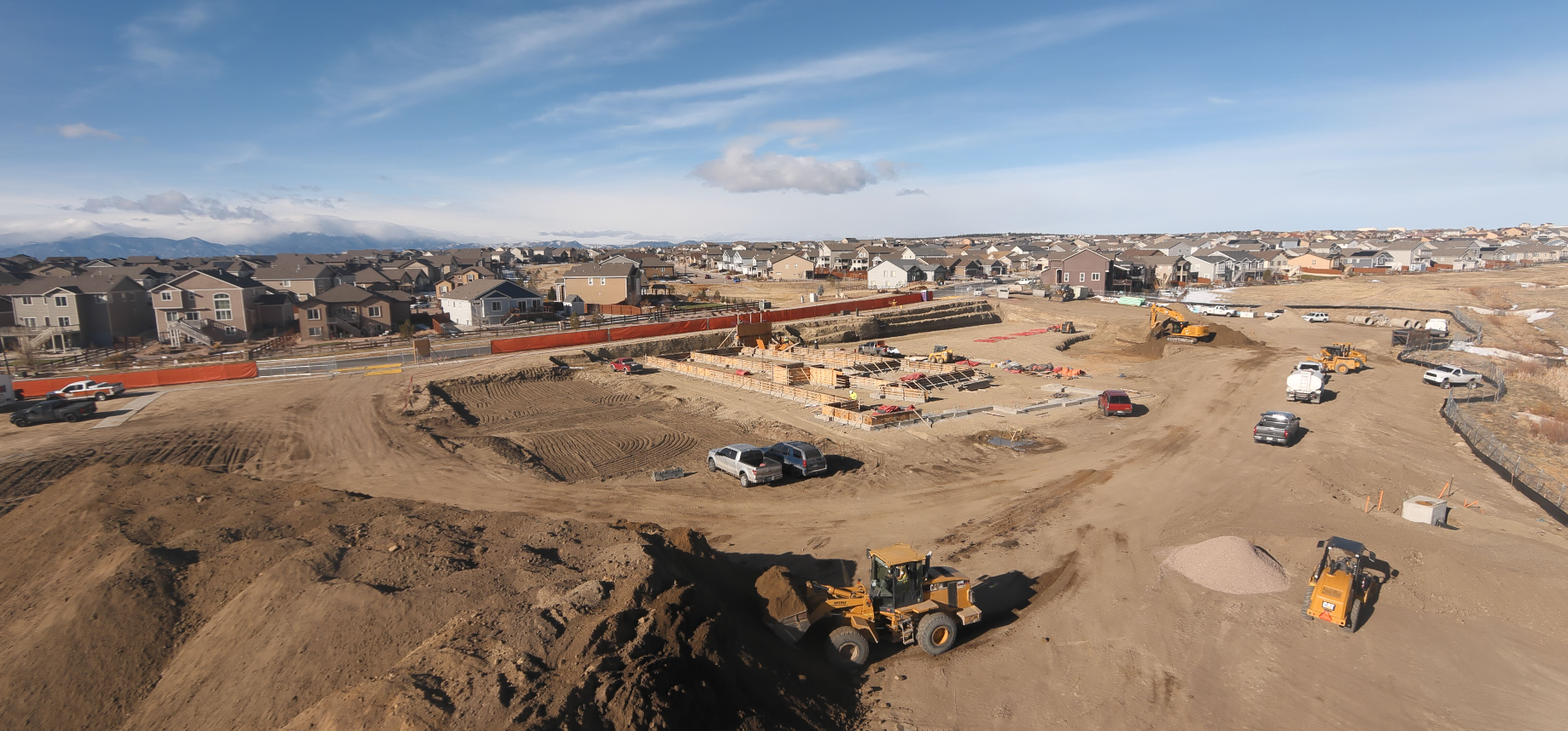 Construction site with machinery and vehicles, surrounded by residential houses under a blue sky.