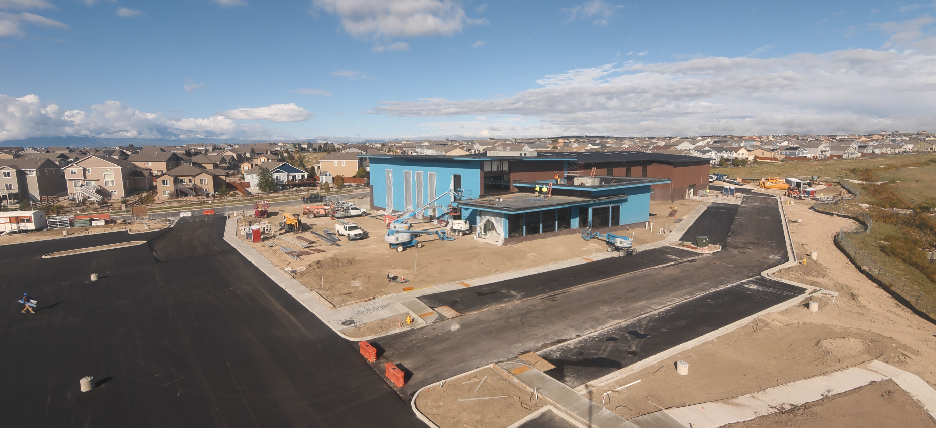 Construction site with a blue building, machinery, and nearby residential area under a clear sky.